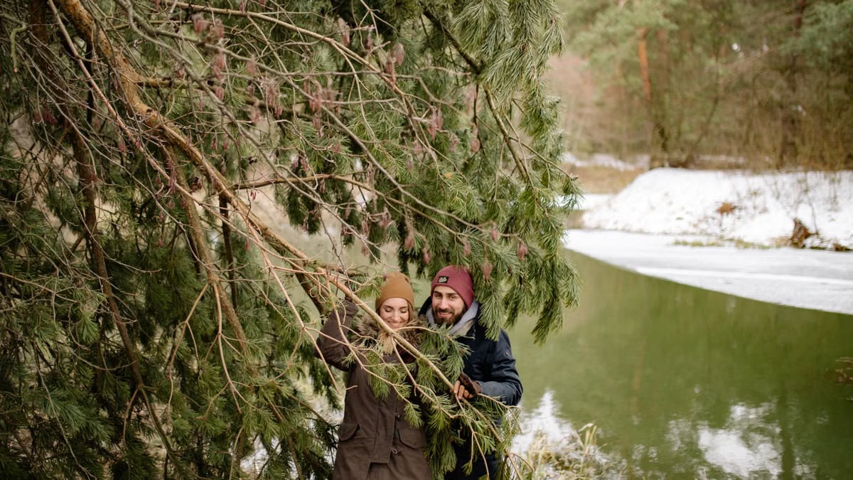 A happy couple exploring a snowy forest, smiling under pine trees by the river.