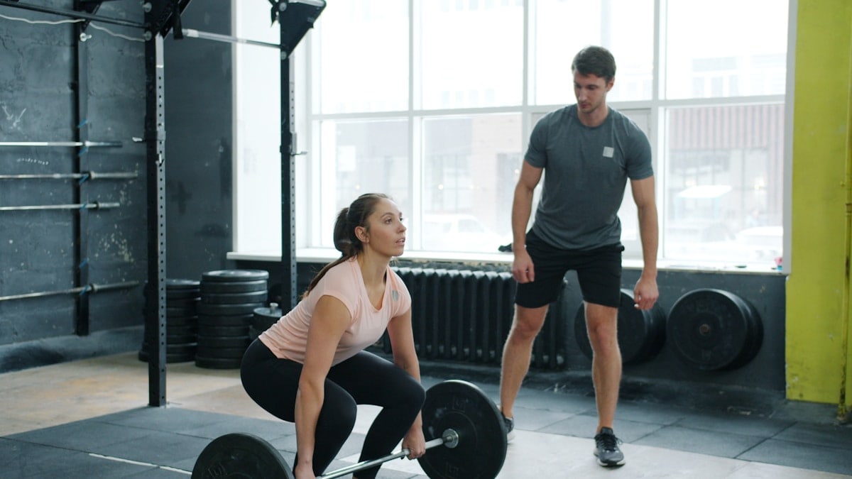 Strong young lady in sportswear is lifting heavy barbell while instructor handsome guy is watching during bodybuilding training in sports gym.